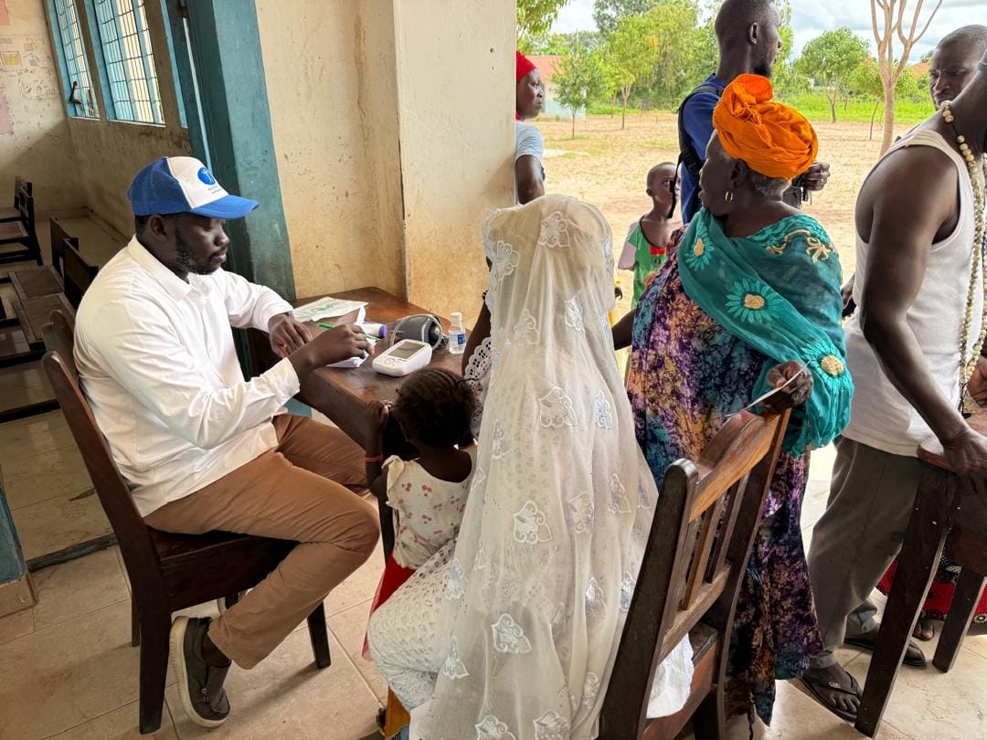 A health worker in a Humanity First cap writes notes while a woman sits with a young child at the consultation desk