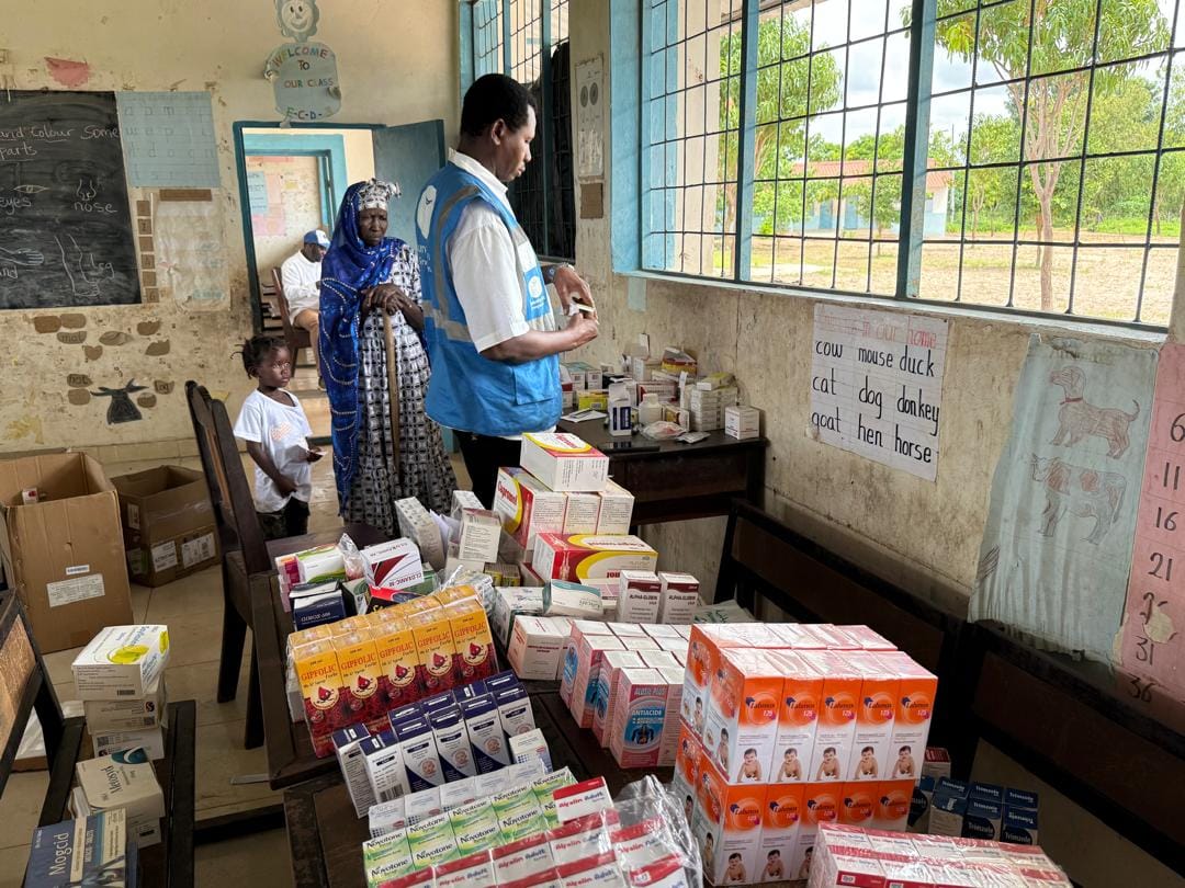 A volunteer in a Humanity First vest sorts medicines laid out on tables while an older woman and a child wait