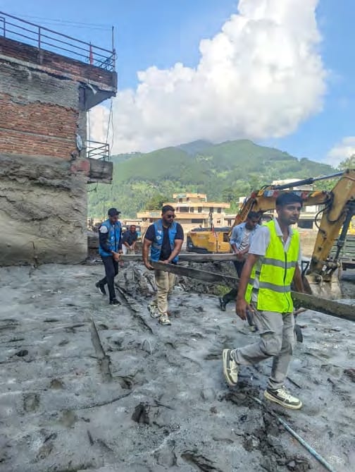 Volunteers in high-visibility vests clear debris beside a damaged building, with an excavator behind them